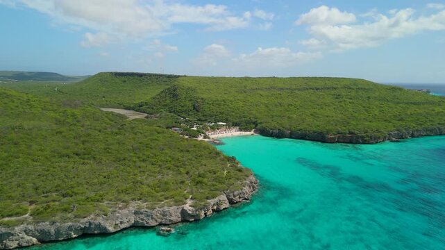 High altitude aerial orbit reveals turquoise bay of Daaibooi Beach nestled between green hills and rocky cliffs in Curacao
