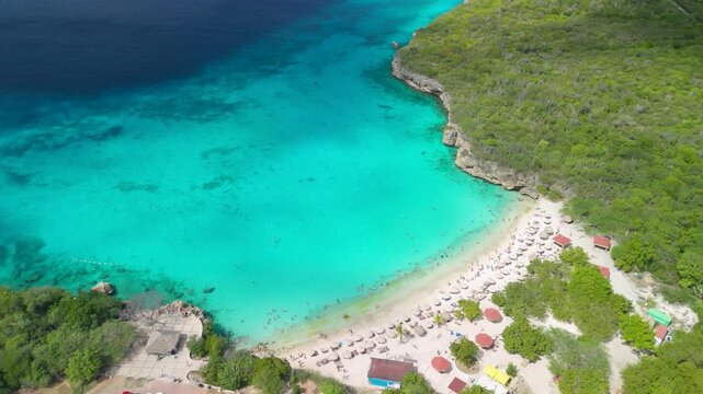 High altitude aerial parallax over Grote Knip beach in Curacao shows turquoise cove, white sand, straw umbrellas and rocky cliffs surrounded by green coastal vegetation