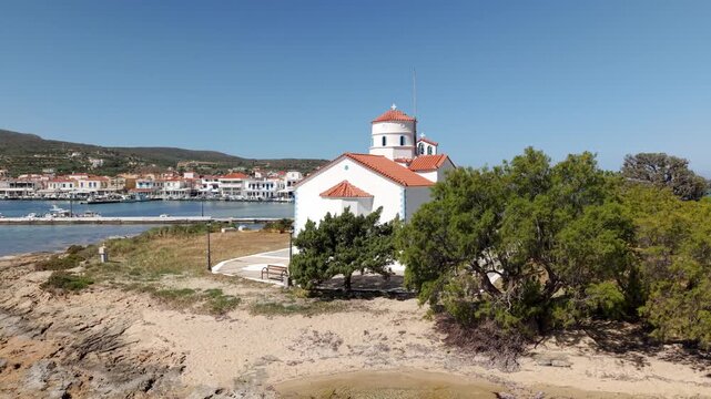 Aerial pullback shot of ancient Agios Spyridon church on small rocky island shoreline on a sunny day, Elafonisos village waterfront houses across the Ionian sea in background