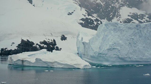 Massive iceberg drifts through Lemaire Channel, Antarctica, with smaller ice floes on calm water and snow-covered rocky mountains in backdrop