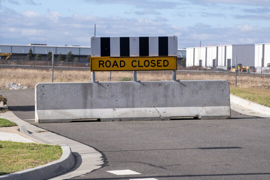A heavy concrete Jersey barrier block with a "ROAD CLOSED"  sign blocks an uncompleted street in Truganina, Melbourne Australia. The roadblock marks a boundary separating an adjacent industrial zone