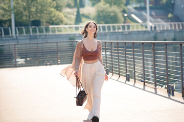 Fototapeta premium Young woman walking along urban waterfront promenade in summer wearing stylish casual outfit and carrying a handbag, smiling confidently under bright sunlight