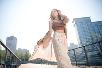 Obraz premium Young woman walking on an urban promenade in summer fashion, smiling and enjoying sunny city life with flowing cardigan and wide pleated pants