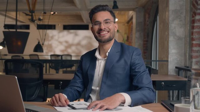 Indian businessman business man signing document looking at camera nod head smiling male entrepreneur with laptop office CEO executive guy corporate paperwork nodding positive answer write in papers