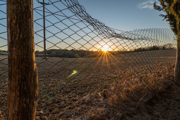 Obraz premium Sunset view over farmland seen through a netted field and wooden fence. The image captures the warm light and open rural atmosphere of Tuscany.