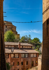 Fototapeta premium View through a narrow opening toward rooftops and greenery in Siena’s historic center. A peaceful composition that mixes old architecture with a natural hillside backdrop.
