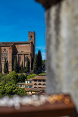 Fototapeta premium View of Siena’s cathedral framed by a blurred foreground element, creating a layered and artistic composition. The city’s historic architecture stands out against the sky.