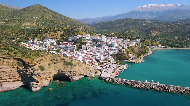Agia Galini Crete Greece Aerial Panorama. White village and orange sandstone cliffs with turquoise sea caves. Snow capped mountains background on Greek island summer travel destination asset