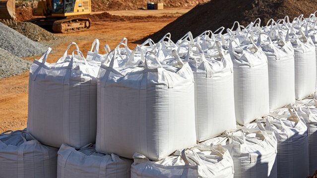Multiple large white bulk bags stacked securely near a construction project with cranes in background.