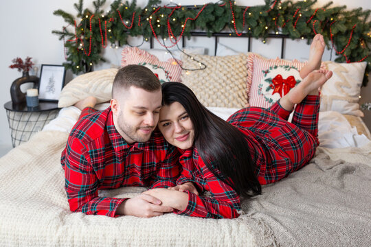 Affectionate young couple in red plaid pajamas lying on bed with feet up during Christmas morning. Concept of shared joy, lifestyle harmony, and a happy modern marriage.