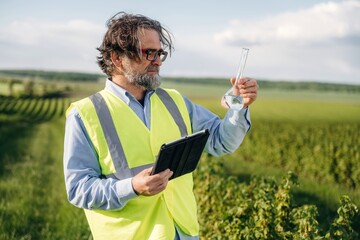 Fototapeta premium Middle-aged man in reflective safety vest examines liquid in a laboratory flask while holding a tablet outdoors in a vineyard landscape under a clear sky