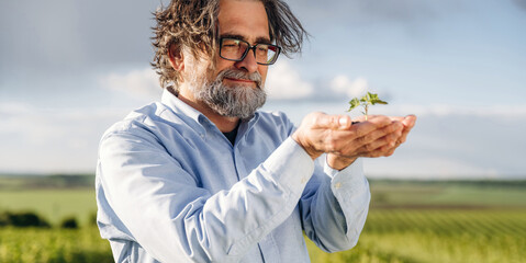 Fototapeta premium Middle-aged man with glasses holding a small green plant in his hands, standing in a lush vineyard landscape under a bright sky with clouds