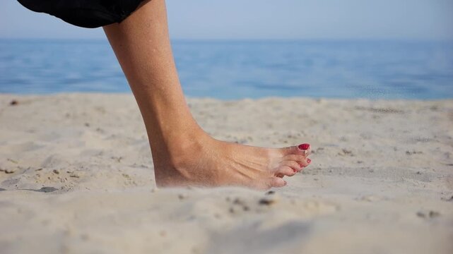 Female feet walking on sand at the beach with seascape at background. Legs of adult woman stepping along sea shore. Barefoot lady strolling near ocean coast. Concept of summer vacation. Low view