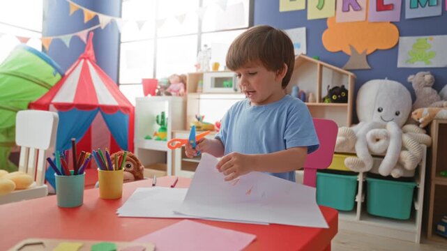 Boy cutting paper with scissors and holding pencil in hands at playroom table in building; concentration.