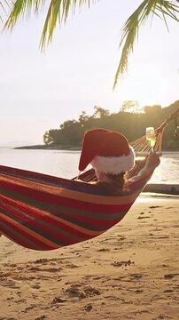 Vertical video. Slow Motion Woman Wearing a Santa Hat Resting in a Hammock While Holding Sparkling Wine Above a Tropical Beach at Sunset, Festive Holiday Relaxation