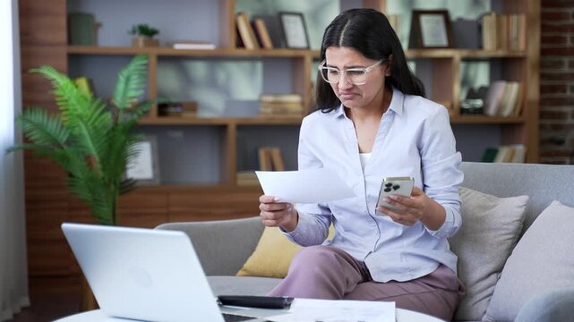 Confused young woman struggling to manage household finances, using mobile phone, sitting on sofa in living room at home office. Puzzled adult female feeling unsure about payments, looking at camera.