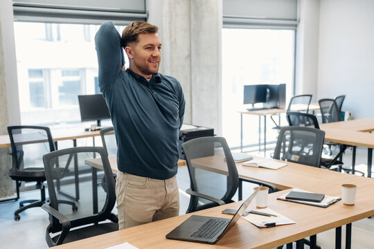 Young Caucasian male stretches while standing at a desk in a modern office environment with empty chairs and computer workstations in the background