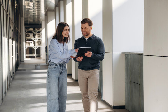 Two professionals, a woman and a man, engaged in discussion while using a tablet in a modern office corridor with large windows and minimalist design elements