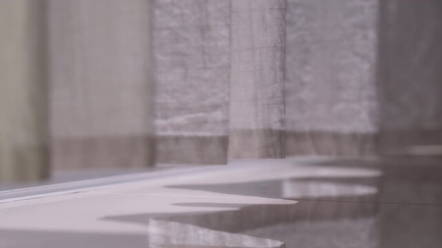 Real time close up of a curtain hem swaying in warm morning light, detailed fabric texture with cozy sun glow leaking underneath onto a reflective room floor.