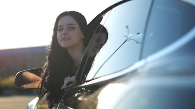 Young beautiful girl leaning out of driver window at modern electric car and looking into camera. Portrait of attractive smiling woman in alternative electrical vehicle. Slow motion