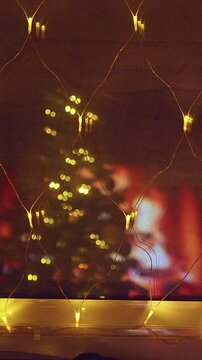 Vertical video. Warm Christmas Window With Golden Fairy Lights Reflecting a Decorated Tree and Fireplace, Cozy Holiday Home, Festive Atmosphere and Winter Comfort