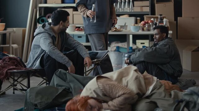 Diverse young adult men sitting in temporary humanitarian shelter while volunteer hands bottle of water to one of them. Clip is perfect for charity and social support campaigns