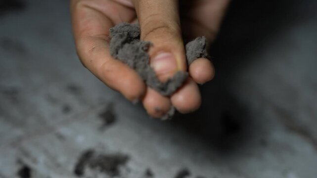 Close-up of a hand holding gray crumbly ash or clay, showing texture and touch in a simple dark setting
