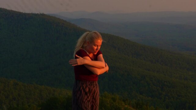 Middle aged blond woman doing breathing exercises practice opening hands outdoors at sunset, scenic mountains background. Attractive active sportswoman making relaxing pranayama for wellbeing, healthy