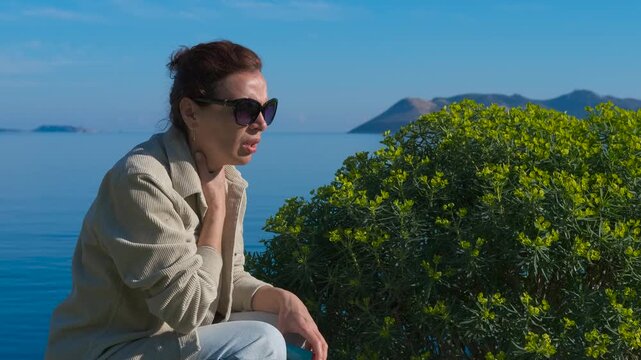 Woman using asthma inhaler near the sea. Mature woman battling asthma symptoms, using medical inhaler while sitting near coastal landscape, breathing deeply and recovering during bright sunny day