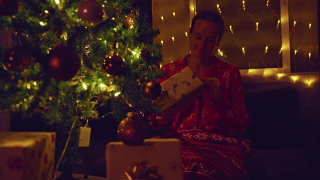 Happy Woman Unwrapping a Christmas Gift Beside a Decorated Tree With Warm Golden Fairy Lights, Cozy Holiday Home, Festive Celebration and Family Tradition