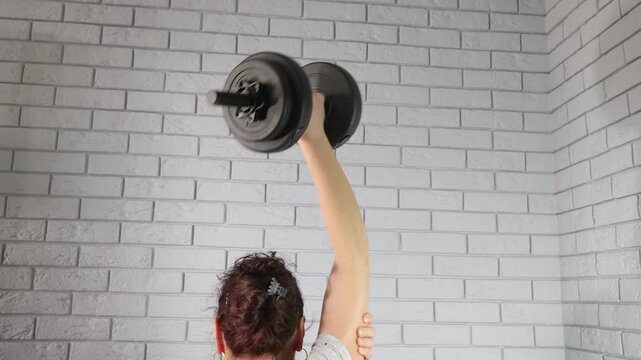Woman performing dumbbell triceps extension exercise. Unrecognizable woman with her back to the camera performing a one-arm overhead dumbbell triceps extension during a home workout