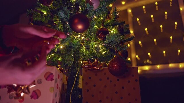 Hands Adjusting Christmas Tree Branches and Ornaments Above Wrapped Gifts With Warm Golden Fairy Lights, Cozy Holiday Home, Festive Decoration and Family Tradition