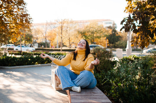 Smiling stylish woman 35-39 year old wearing knitted sweater sitting on bench at city street over yellow leaves. Autumn season.