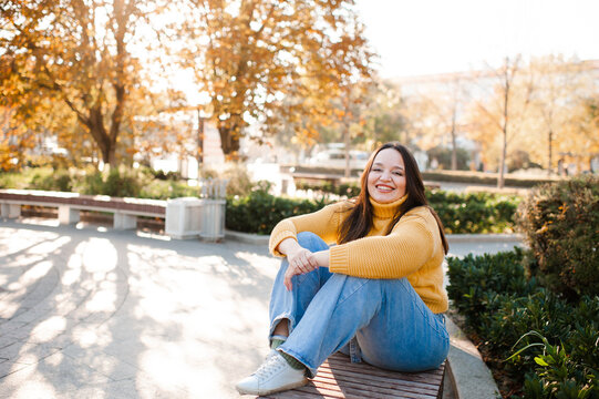 Smiling stylish woman 35-39 year old wearing knitted sweater sitting on bench at city street over yellow leaves. Autumn season.