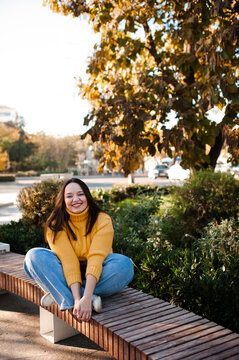 Smiling stylish woman 35-39 year old wearing knitted sweater sitting on bench at city street over yellow leaves. Autumn season.