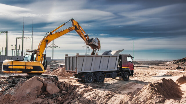 A dump truck gets loaded by an excavator at a construction site. The work takes place during daylight under a clear sky with power lines visible
