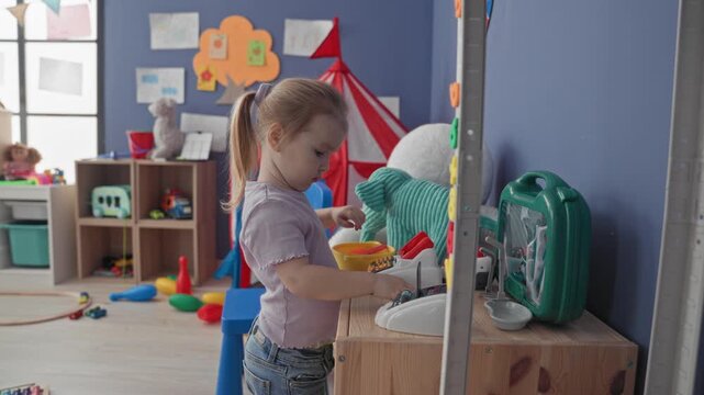Girl pressing buttons on a toy cash register in a bright playroom with toys and storage shelves; concentration imagination.
