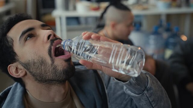 Closeup of Middle Eastern young adult man drinking water from plastic bottle during humanitarian aid distribution. Ideal for nonprofit campaigns, disaster relief, and clean water advocacy videos