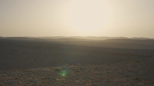 Sunset glows over the vast desert landscape. Low hills fade into the hazy horizon. Green lens flare appears in the foreground. Empty sandy terrain stretches to the evening sun. Ramon Crater in Israel