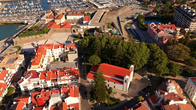 A drone reveals a white church surrounded by a red roofed coastal town near a marina
