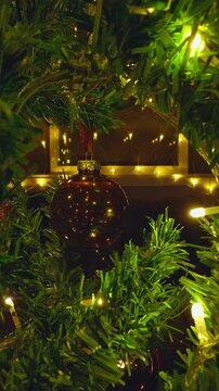Vertical video. Christmas Tree Branches and Glowing Baubles Framing Warm Window Lights and Cozy Holiday Interior in a Creative Point of View Shot