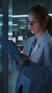 Vertical Screen. Concentrated Caucasian Female Scientist Uses Digital Tablet Beside Wall Display in Medical Laboratory. Physician Analyzes Data for Drug Development and Disease Research.