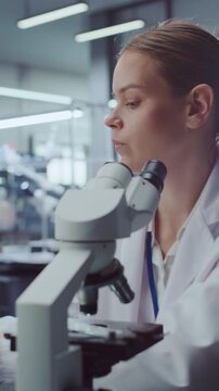 Vertical Screen. Female Scientist Examines Sample Through Optical Microscope, Then Looks Aside Thoughtfully in Modern Medical Research Laboratory. Physician Conducts Analysis for Drug Development.
