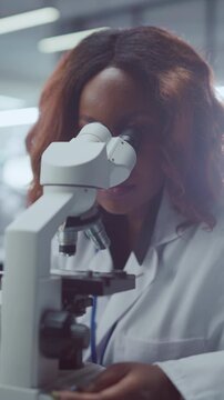 Vertical Screen. Diligent Black Female Scientist Looks Into Optical Microscope Eyepiece, Carefully Inspecting Biological Specimen in Modern Medical Laboratory. Researcher Develops Innovative Treatment