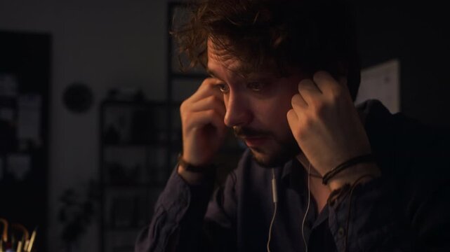 Caucasian young adult man putting on earphones and starting online video call on laptop while working late in office. Clip is suitable for remote work and corporate communication videos