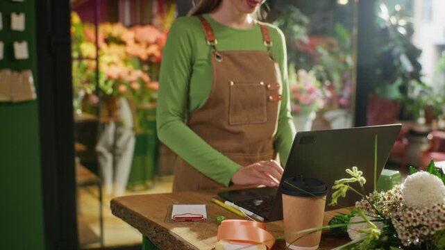 Young adult Caucasian female florist typing on laptop and writing notes on notepad in flower shop. Clip is ideal for small business management and e-commerce marketing campaigns