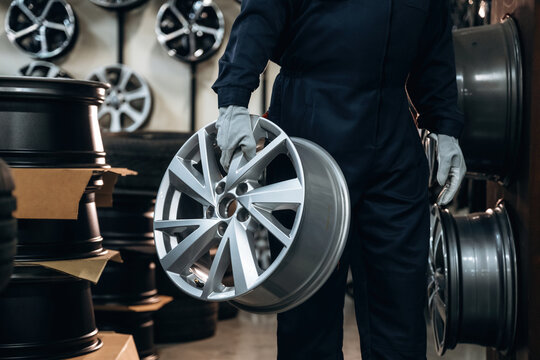Male technician in dark coveralls holds a silver alloy wheel while surrounded by stacks of tires and additional wheels in a well-lit automotive workshop