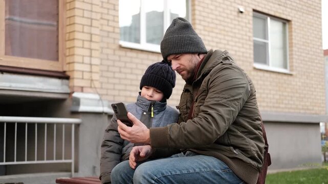 Caring father showing smartphone screen to little son outdoors. Dad and boy sitting near house, using mobile phone together. Digital Parenting, Screen Time Management. Parent and child with smartphone