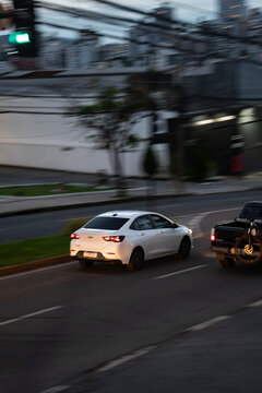 White Chevrolet Onix Plus rear view cruising city street, Subcompact sedan on urban traffic at twilight
