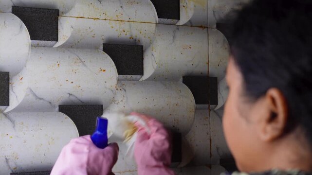 A woman wearing pink gloves scrubbing greasy kitchen wall with microfiber and chemical spray, hygiene and ensuring spotless kitchen cleaning maintenance concept.